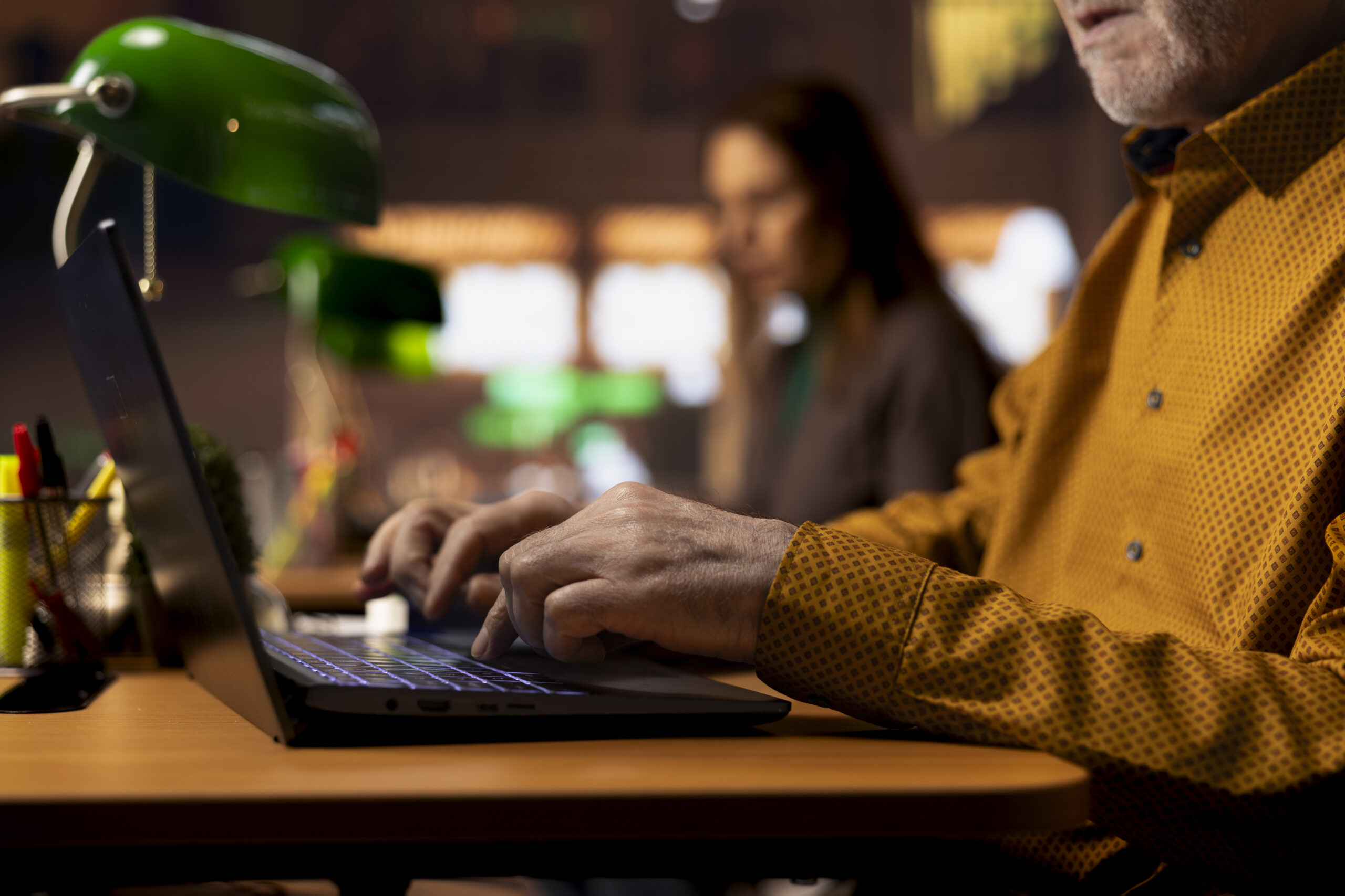 An older man typing on a laptop in the foreground with a green lamp.