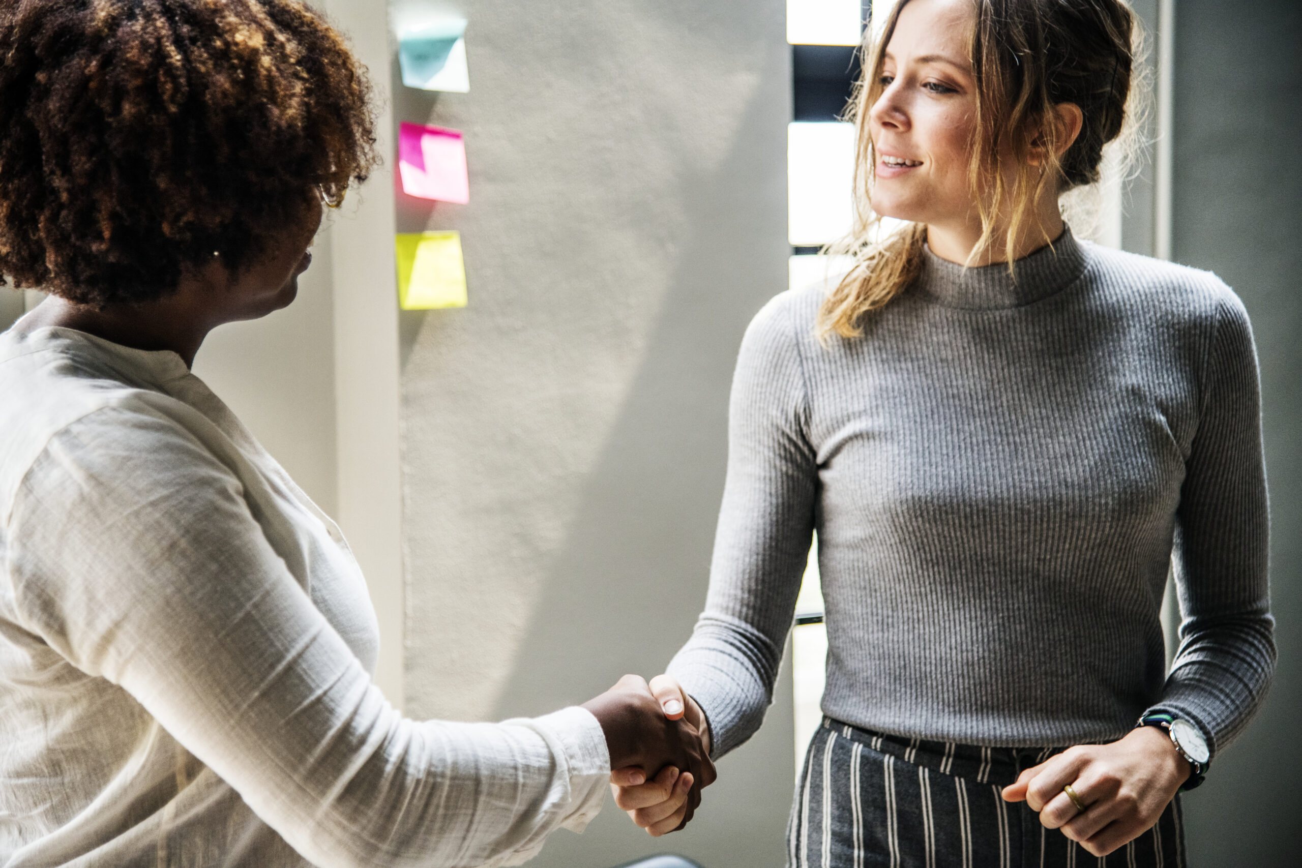 Two business women shaking hands in a bright office space, standing near a wall with colourful sticky notes.