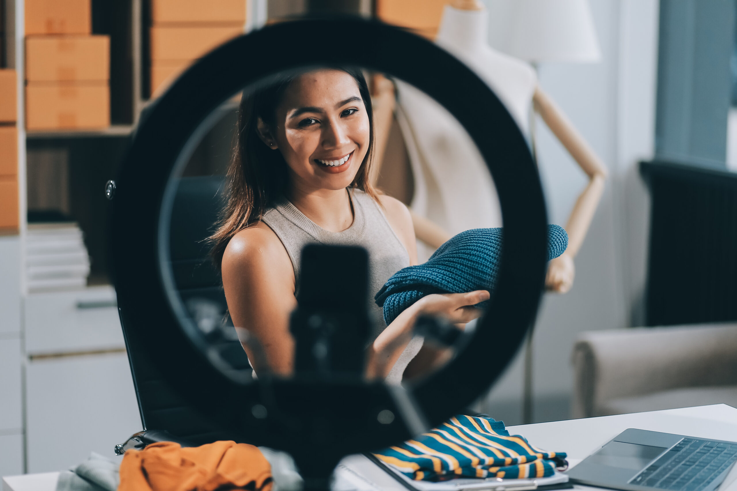 a micro influencer recording a video. in the foreground there is a ring light stand with a phone and the influencer is in the background holding clothes.