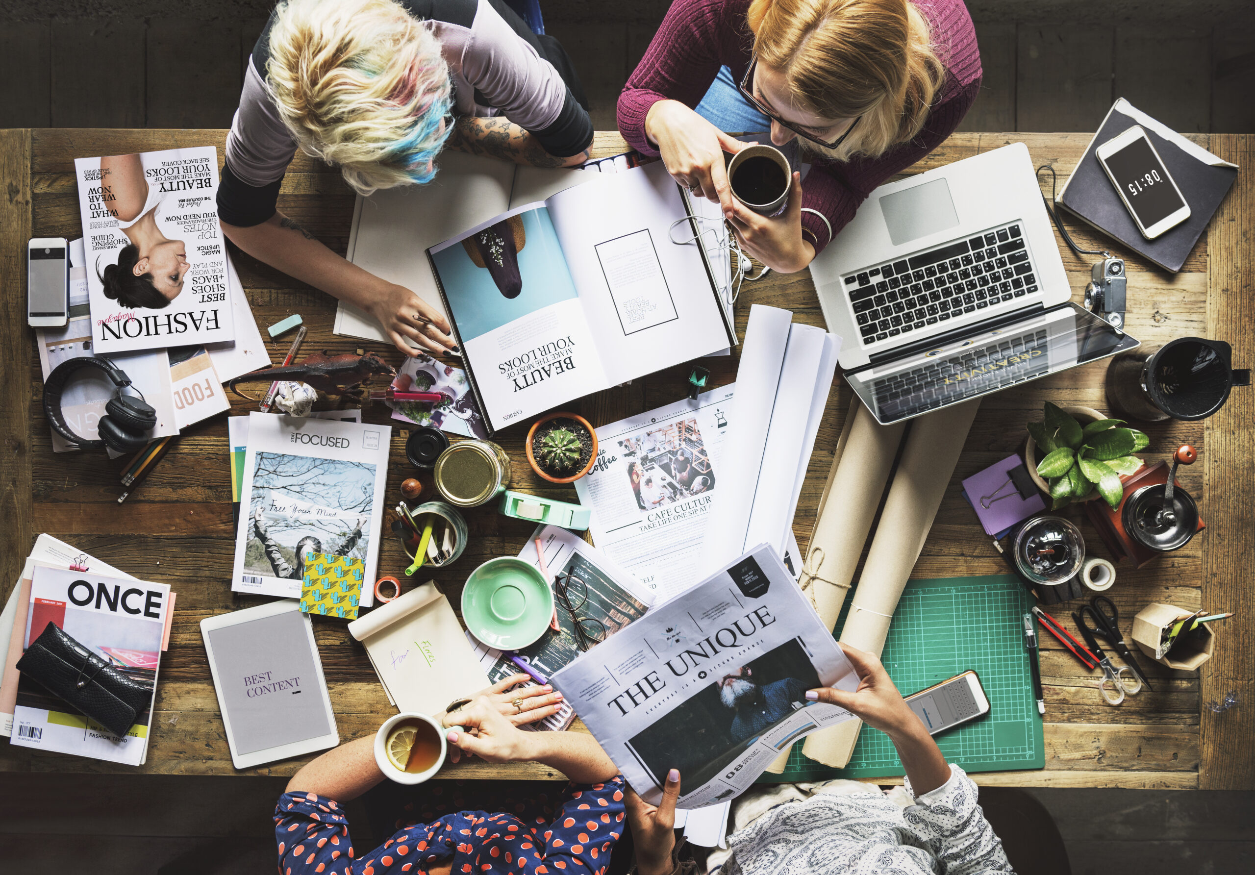 Creative team working on media planning and buying strategies at a shared desk with magazines, newspapers, laptop, coffee cups, and marketing materials.