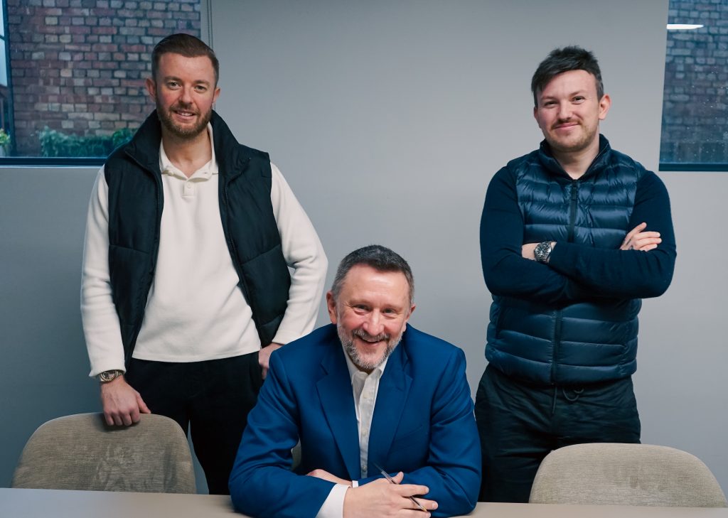 Three men are posing indoors in a modern office setting. The man in the middle (David) is seated at a table wearing a blue suit and smiling, while the two men beside him (Scott, left, Jamie, right, of Podcast-Room) are standing and wearing casual padded vests. Two windows and a brick wall outside are visible in the background.