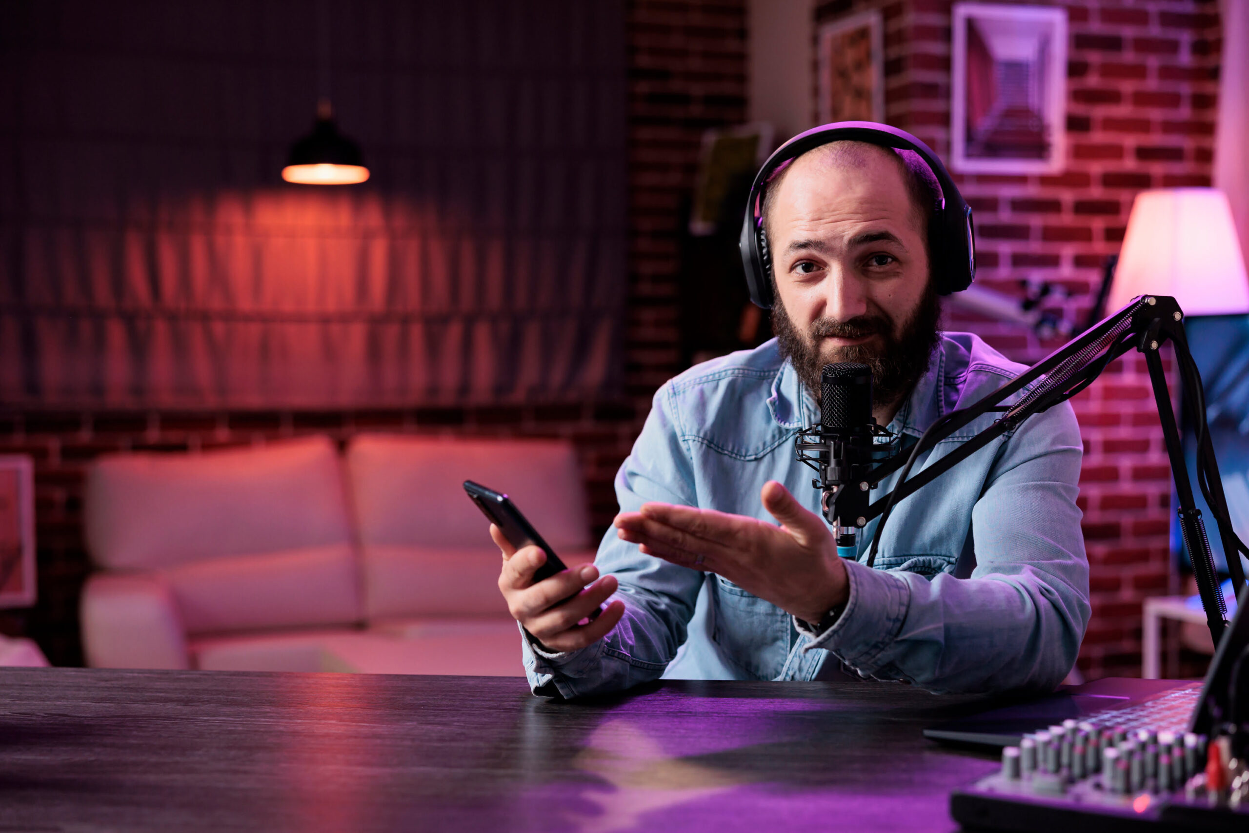 A bearded man wearing headphones sits at a podcast desk with a microphone, holding a smartphone and gesturing as he speaks. The background features warm studio lighting, a sofa and exposed brick walls, creating a casual podcast recording setup.