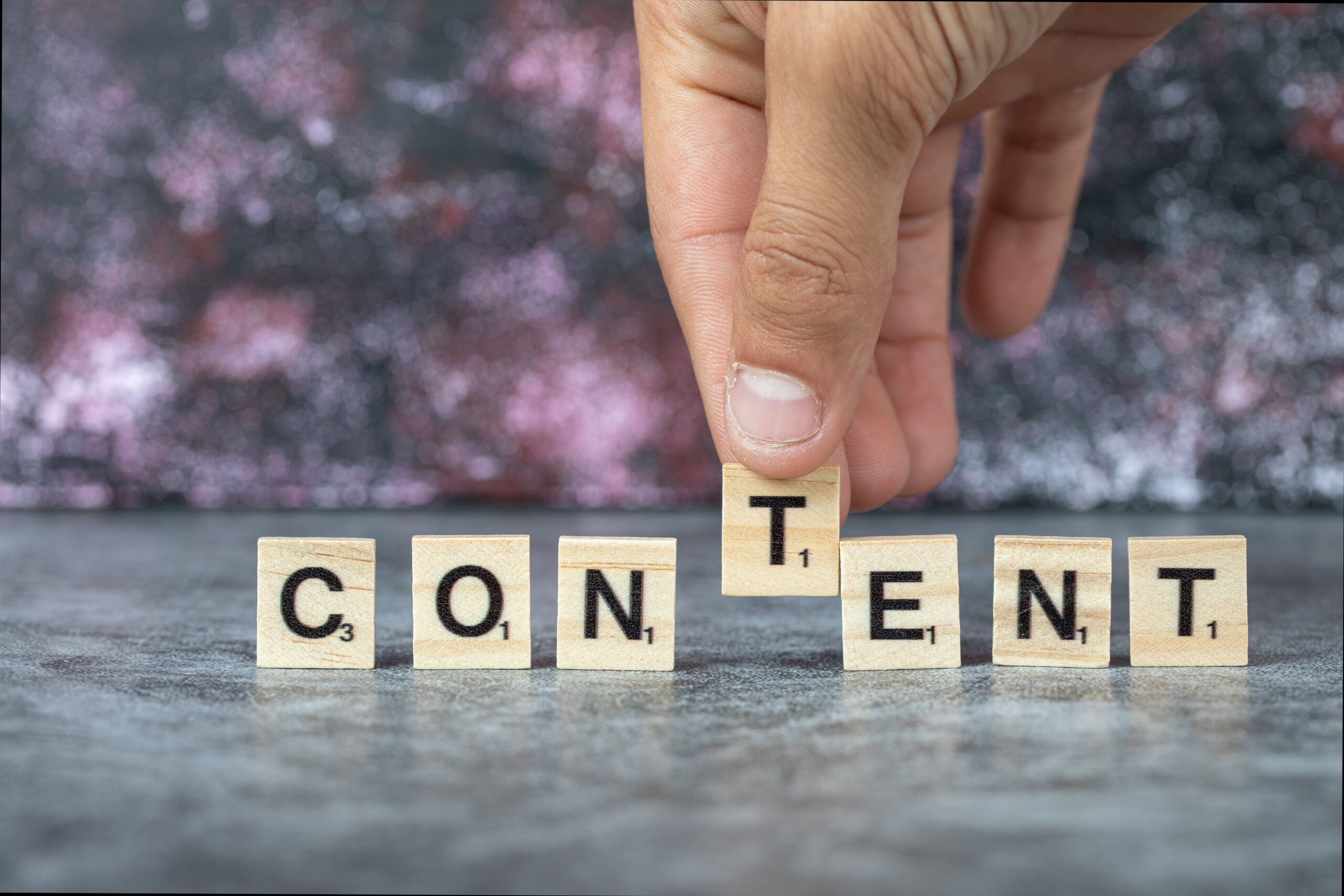 Hand placing a wooden letter block to complete the word “CONTENT” spelled out on small wooden tiles against a blurred background.