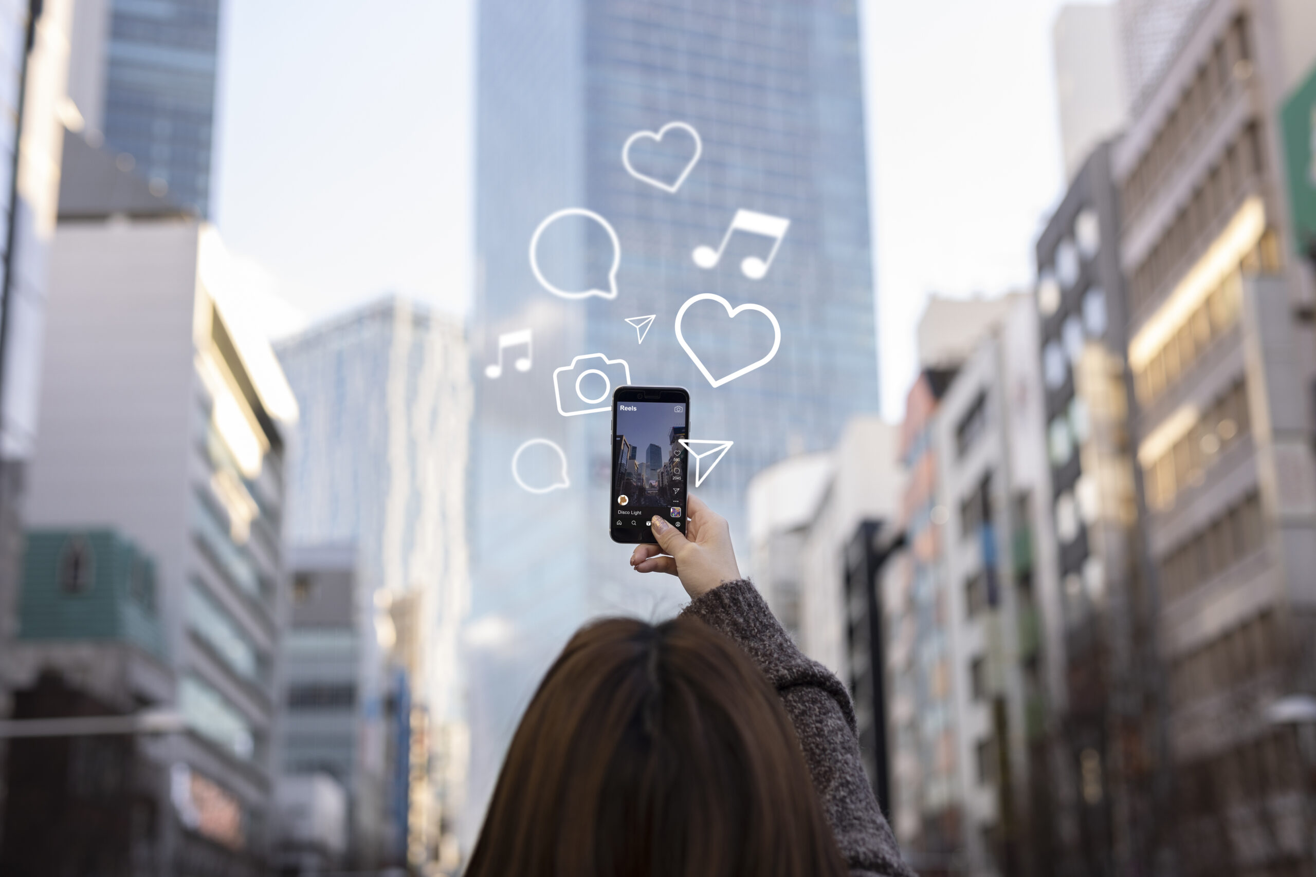 Person holding a smartphone filming a city street with illustrated social media icons including hearts, speech bubbles, a camera, and music notes floating above the screen.